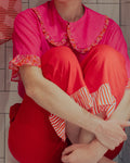 Une chemise rose fuschia à maxi col claudine et manches volantées et un pantalon rouge à bas volantés rayés sont portés par une femme assise en tailleur